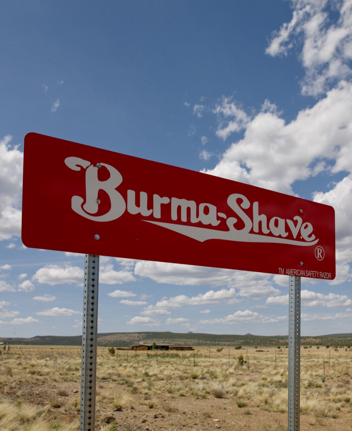 A red Burma-Shave sign on metal posts in front of open Arizona grassland and a big sky