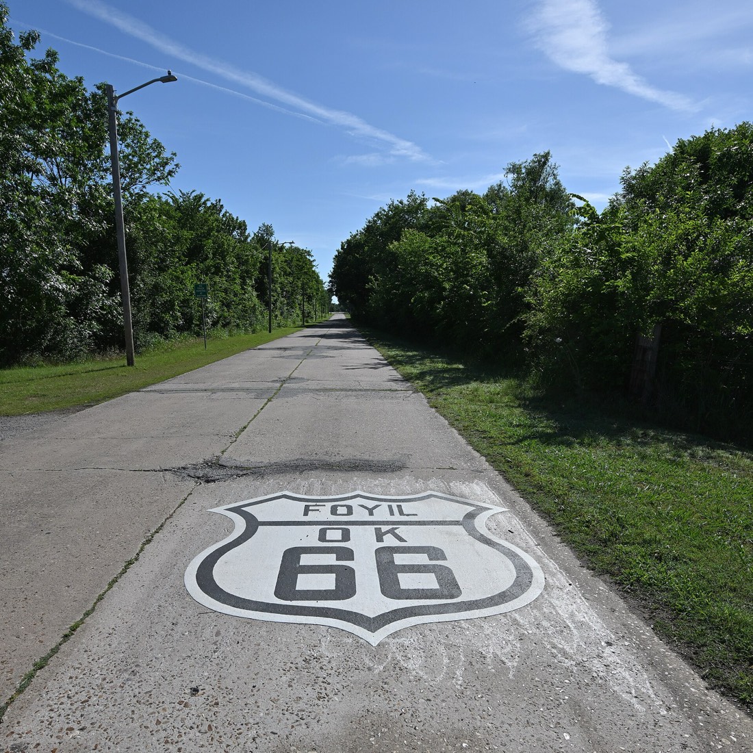 A painted Foyil OK 66 shield on an old concrete road lined with trees, under a blue sky