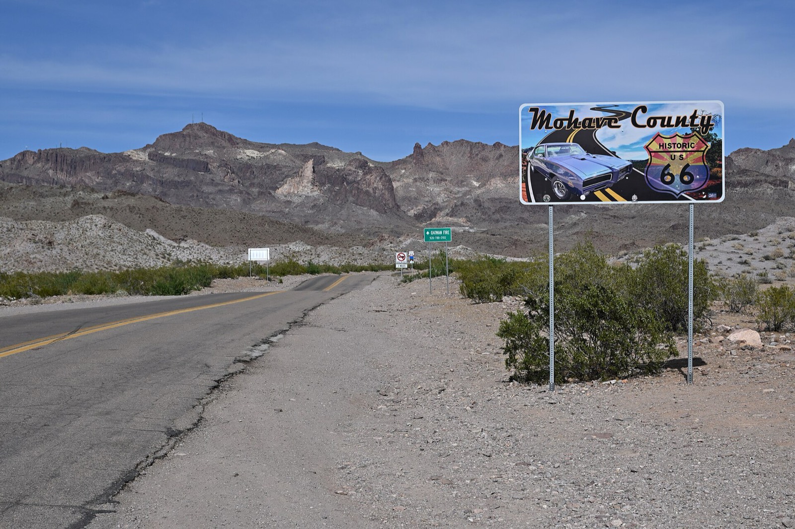 A Mohave County Historic US 66 sign beside the two-lane Oatman Highway, with desert scrub and jagged mountains behind