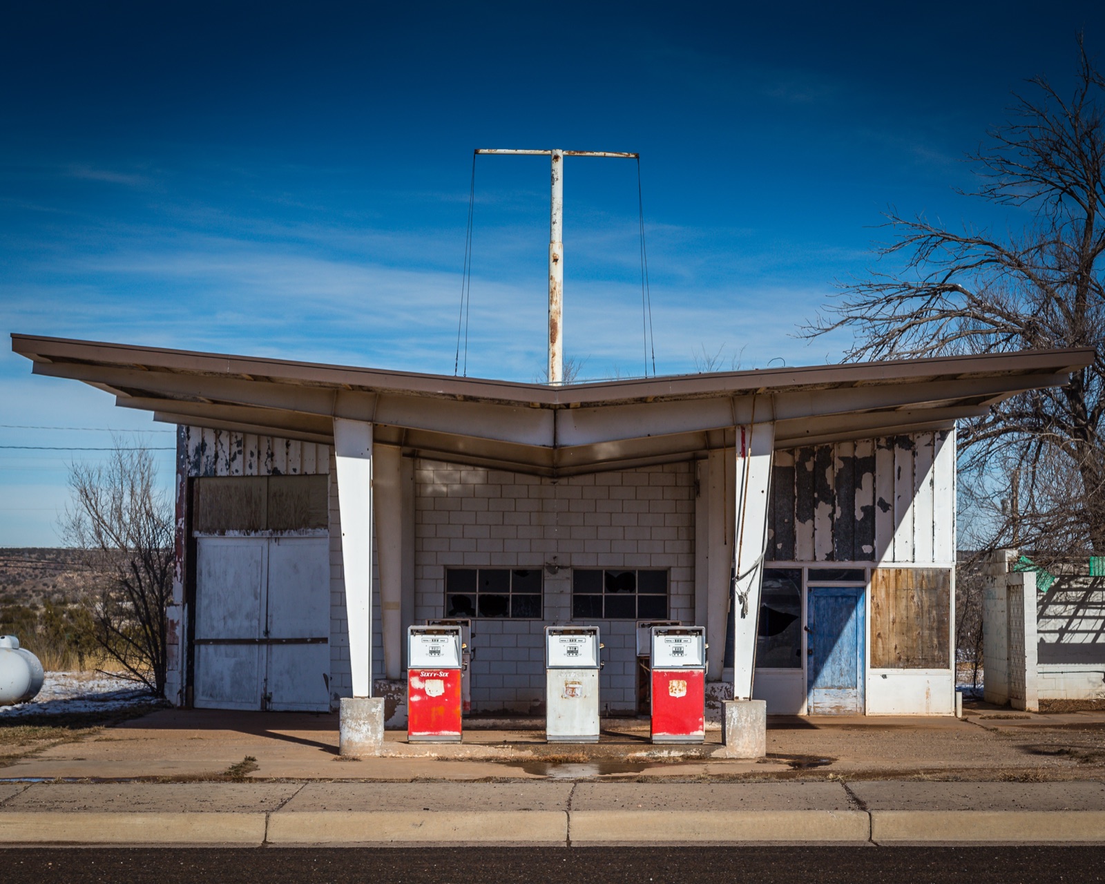 Closed mid-century gas station with old red and white pumps under a swept-wing canopy, patches of snow on the ground and bare trees, in Santa Rosa, New Mexico