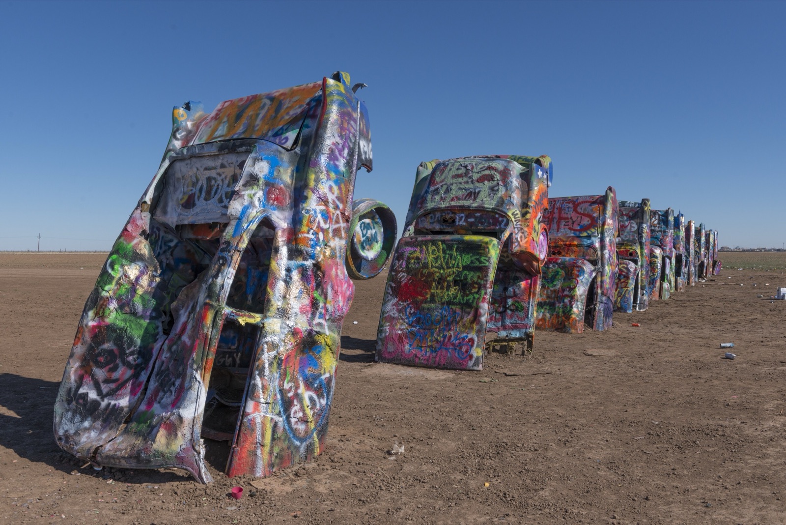 A row of graffiti-covered Cadillacs buried nose-down in a dirt field at Cadillac Ranch, west of Amarillo, Texas, under a blue sky