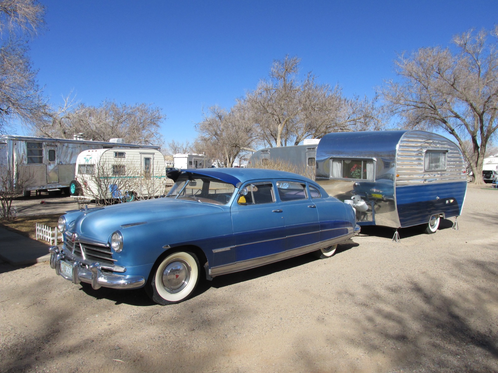 A blue 1950s Hudson Commodore towing a polished vintage camper trailer, with more vintage trailers parked behind, at Enchanted Trails in Albuquerque
