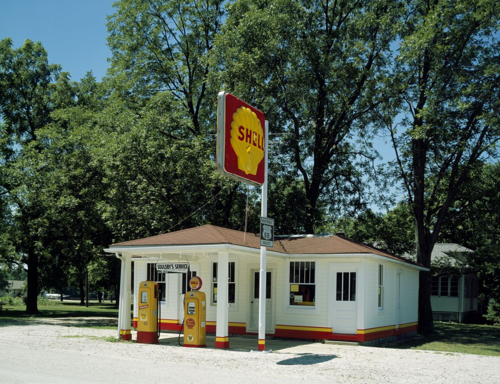 The restored 1926 Soulsby Shell station in Mount Olive, Illinois, a small white building with red and yellow trim, two vintage Shell pumps and a tall Shell sign under big shade trees
