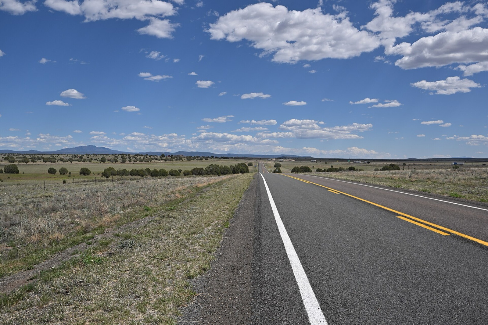 Empty two-lane Route 66 running straight through open grassland toward distant mountains on Crookton Road, Arizona, under a blue sky with white clouds