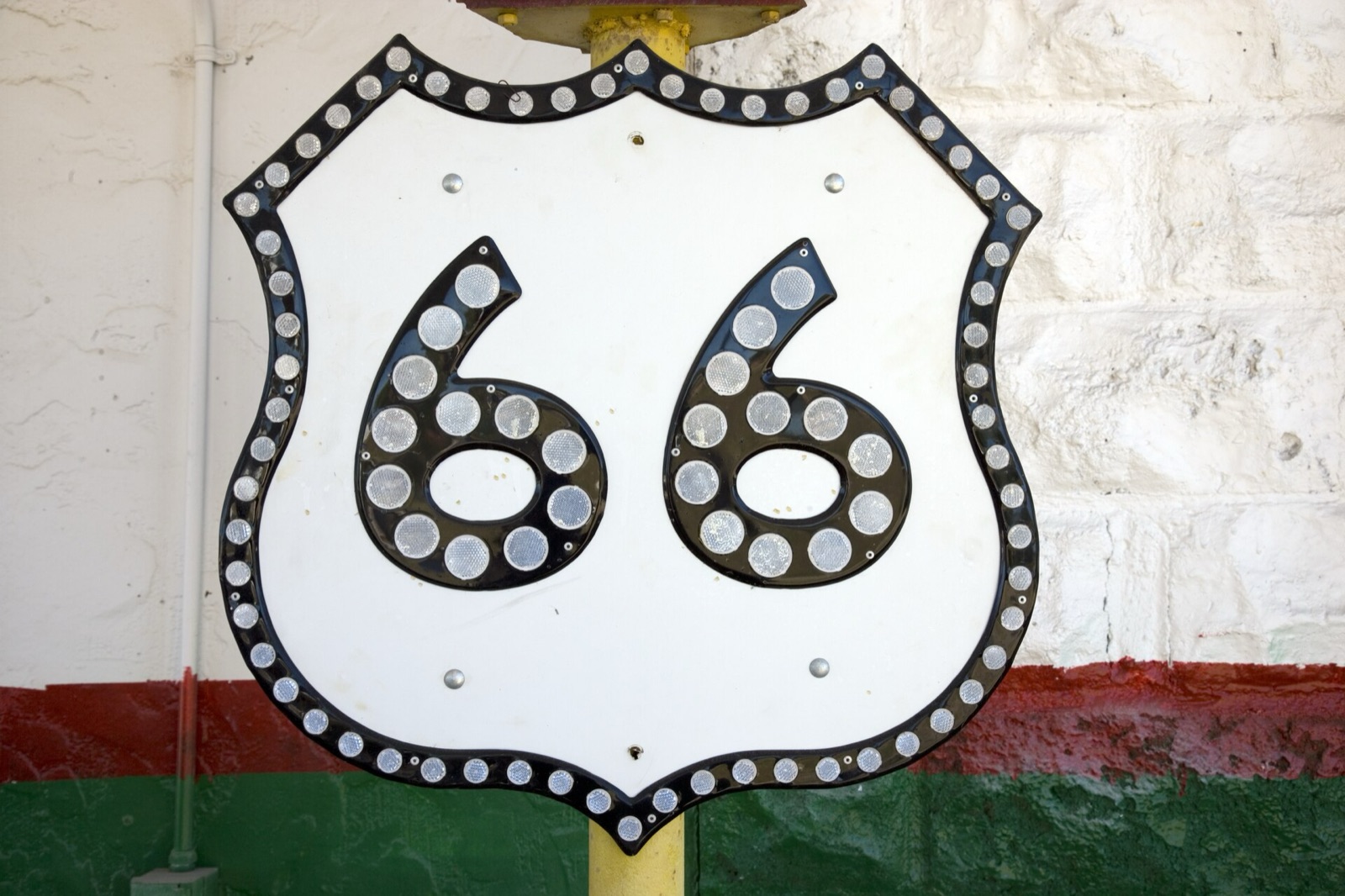A vintage Route 66 shield sign studded with reflectors, mounted on a yellow post against a white wall