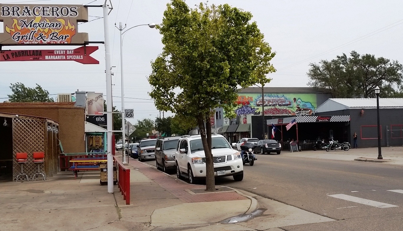 Sixth Avenue in Amarillo, Texas, with parked cars and motorcycles, a Route 66 shield on a lamppost, a restaurant sign and a mural on a low storefront