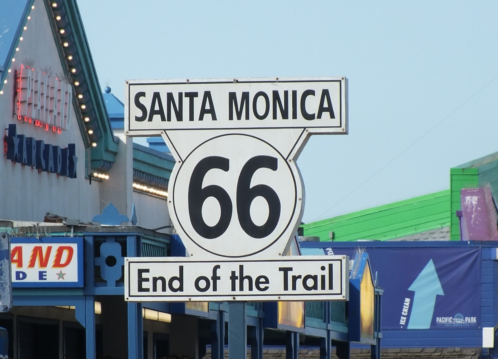 Black and white Santa Monica 66 End of the Trail sign on the Santa Monica Pier, with pier buildings behind it under a blue sky